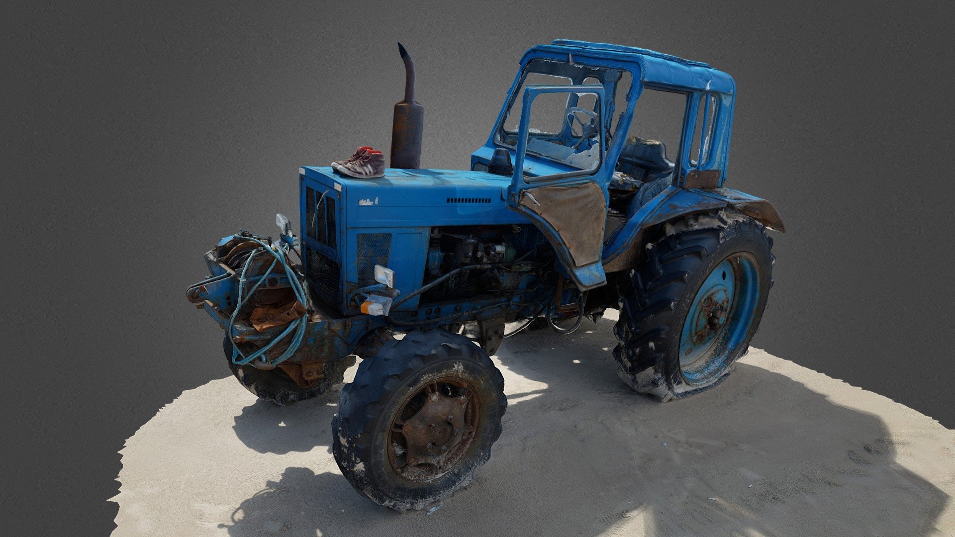 Old, rusty, blue tractor seemingly abandoned at a sea shore. 
Rusty metal, one flat tire, sand on tires, broken glass. 
A pair of sneakers on the front for some reason.